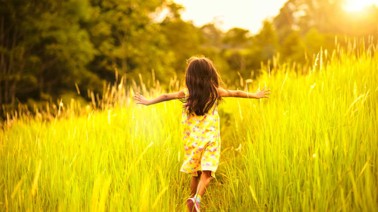 A young girl wearing a yellow sundress with pink flowers runs through a field of tall grass with her arms out.
